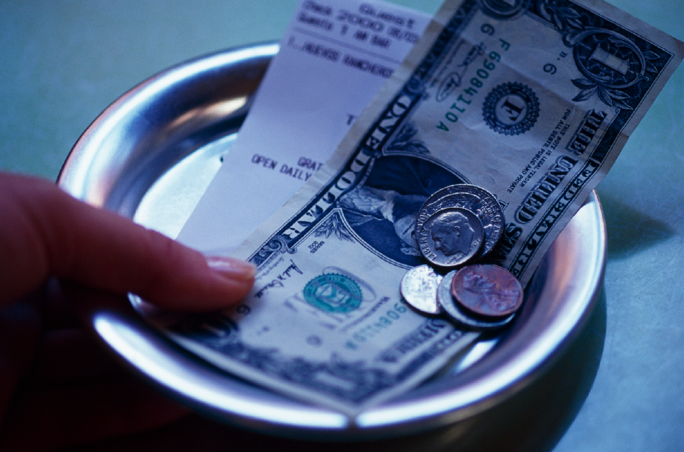 A tipped worker's hand collecting a tip consisting of a dollar bill and some coins on a small white plate.