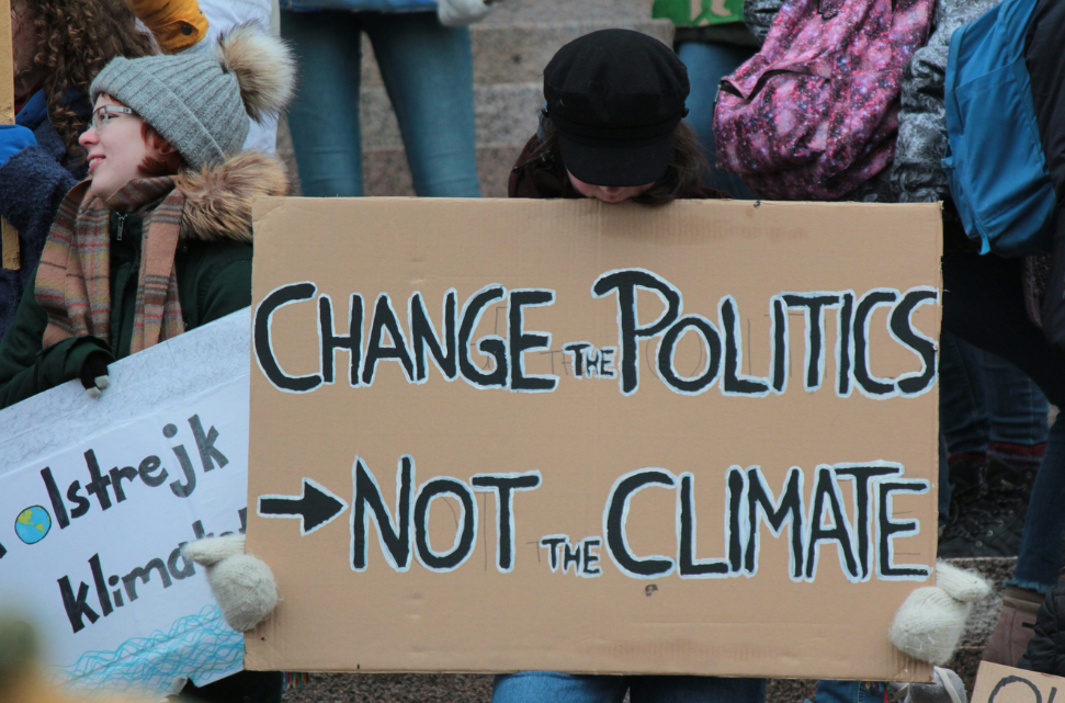 Rising Cost of Debt: An Obstacle to Achieving Climate and Development Goals. A person holding a large cardboard sign that reads "change the politics, not the climate" at a climate change protest, surrounded by other demonstrators.
