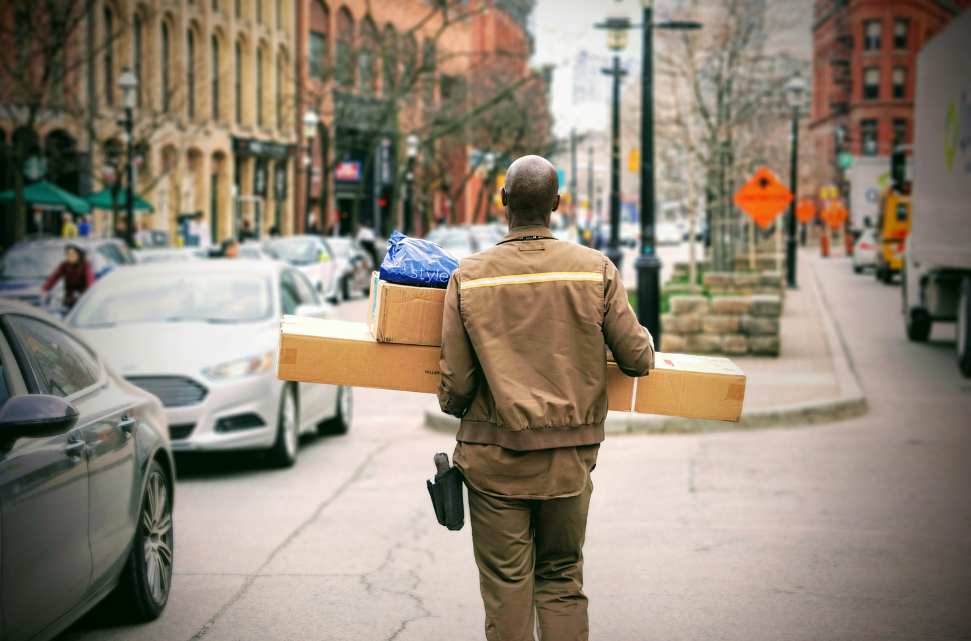 A delivery man in a brown uniform carrying boxes and a smaller blue-bagged item on a bustling city street symbolizing job growth.