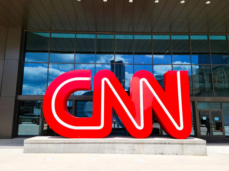 Large red cnn logo in front of a modern building with glass facade and clear blue skies in the background.