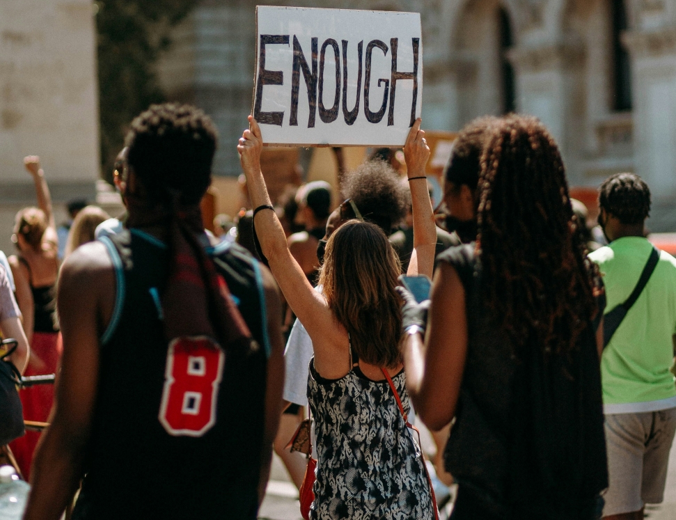 A diverse group of students at a protest against student debt with one individual prominently holding up a sign that reads "ENOUGH" against a sunny, outdoor background.