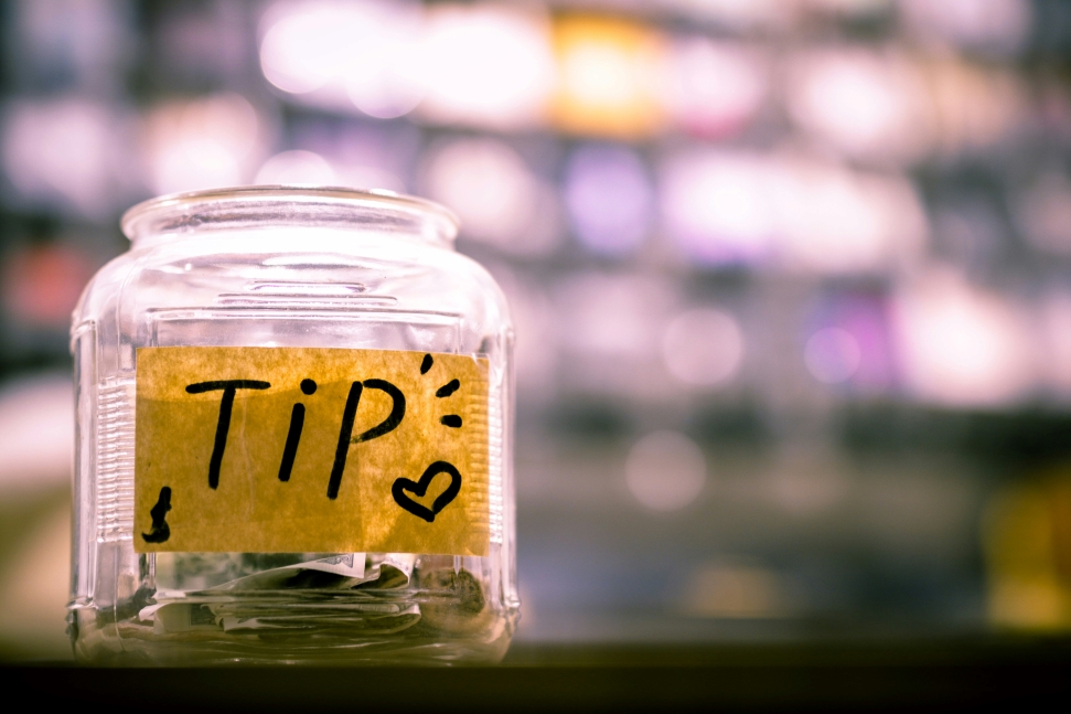 A glass tip jar with paper currency and coins inside is placed on a counter, indicating gratuities. The jar has a hand-drawn label with the word "TIP" and a small heart. The background is blurred with soft, colorful lights.